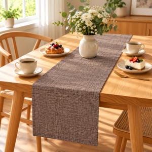 May include: A rectangular dark brown table runner with a woven fabric texture laid across a light wood dining table. The runner features a subtle cross-hatch pattern and is centered beneath a white vase filled with white flowers and greenery.