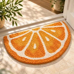 Peut inclure: Un tapis en forme de demi-cercle représentant une tranche d'orange avec des segments texturés orange et jaune séparés par d'épaisses bordures blanches. Le tapis présente une surface touffetée et moelleuse, imitant l'aspect d'un agrume.