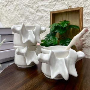 Puede incluir: Tres tazas de cerámica blanca con asas esculturales de formas orgánicas e irregulares. Una taza incluye un asa única con forma de dedo humano apuntando hacia arriba, con detalles sutiles en tonos rosados en la uña y el nudillo.