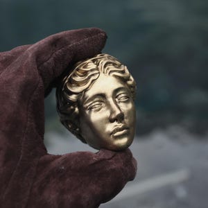 Peut inclure: Une sculpture de tête humaine de couleur or métallique présentant des traits du visage détaillés et des cheveux ondulés. La sculpture est réalisée dans une finition dorée monochrome avec une surface lisse, capturant l'apparence d'un buste classique.
