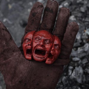Puede incluir: Una escultura de color rojo oscuro que presenta tres rostros humanos interconectados con bocas abiertas y rasgos expresivos. La escultura está elaborada con un acabado suave y pulido, mostrando detalles intrincados alrededor de los ojos, narices y bocas.