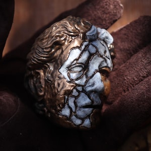 Puede incluir: Una figura de cabeza esculpida con un acabado de dos tonos. Un lado del rostro está representado en un color bronce metálico con textura de cabello detallada, mientras que el otro lado muestra una textura agrietada de color azul claro y blanco.