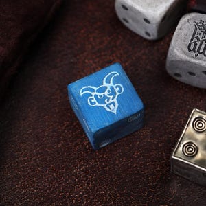 May include: A blue six-sided die featuring a white engraved graphic of a horned creature on its top face. The die is positioned alongside other dice, including a weathered grey die with black pips and a metallic silver die with circular patterns.