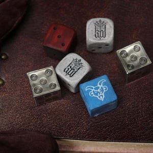 May include: A collection of five six-sided dice featuring varied designs. Two are metallic silver with circular pips, two are light gray stone-like material with a crown emblem, one is dark red wood, and one is blue with a white goat head graphic.