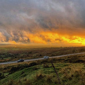 May include: A landscape view of a road cutting through a grassy, rolling countryside under a dramatic sky. Dark, heavy storm clouds contrast with a bright, golden sunset glowing on the horizon, illuminating the hills and two cars on the road.