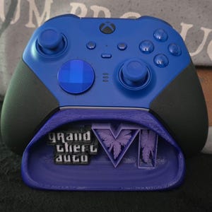 May include: A blue Xbox wireless controller with black textured grips resting on a purple 3D-printed stand. The stand features the text Grand Theft Auto and the Roman numeral VI with palm tree silhouettes integrated into the design.