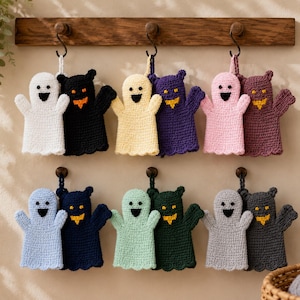 May include: A collection of twelve crocheted ghost-shaped washcloths or hand puppets arranged in pairs. Each pair features one light-colored ghost with a black smile and one dark-colored ghost with orange eyes and a jagged orange mouth.