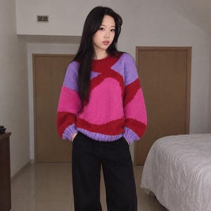 Peut inclure: Un pull en tricot à manches longues présentant un motif color-block audacieux dans des tons vifs de magenta, rouge profond et violet clair. Le pull possède une encolure ronde, des poignets et un ourlet côtelés, porté avec un pantalon noir.