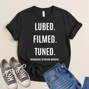 May include: A black short-sleeve t-shirt featuring white text that reads LUBED. FILMED. TUNED. MECHANICAL KEYBOARD MODDING. The shirt is displayed alongside a pair of grey canvas sneakers and blue denim jeans, showcasing a casual apparel set.