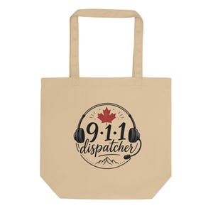 May include: A beige tote bag featuring a circular graphic with black headphones, a red maple leaf, and the text 9.1.1 dispatcher written in a stylized font. Below the text is a black mountain range illustration within the circular border.