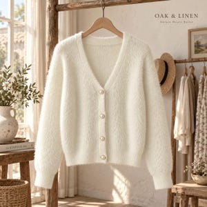 May include: A fuzzy, off-white V-neck cardigan featuring long sleeves, ribbed cuffs, and four round pearl-like buttons down the front. The text OAK & LINEN Nature Wears Better is visible in the upper right corner of the image.