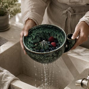 May include: A dark green and black speckled ceramic citrus juicer bowl with a central reamer and a side handle. The bowl contains fresh blueberries and raspberries, with water draining through the base into a sink below the ceramic vessel.