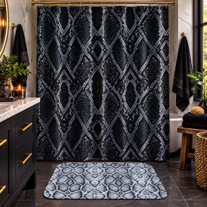 May include: A shower curtain and a matching rectangular bath mat, both featuring a repeating black and grey snakeskin pattern. The shower curtain hangs from a gold rod with gold rings, while the bath mat lies flat on the dark tiled floor.