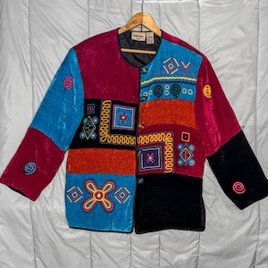 Puede incluir: Una chaqueta de manga larga con un diseño de parches de terciopelo en rojo, azul, naranja y negro. La prenda está adornada con coloridas formas geométricas, espirales y líneas bordadas, e incluye un cierre frontal de botones.
