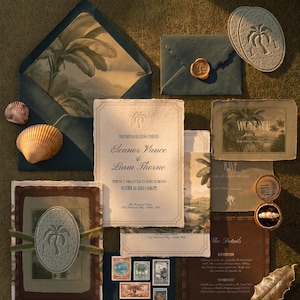 May include: A wedding stationery suite featuring deckled-edge paper, dark blue envelopes with tropical interior liners, vintage postage stamps, a gold wax seal, two seashells, gold leaf accents, and a small ring box containing a pearl ring.