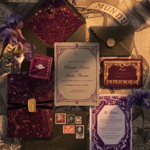 May include: A wedding invitation suite featuring a cream-colored main card with purple ornate borders, a dark purple celestial-themed envelope, a velvet ring box with a gold ring, an RSVP card, a details card, and a black envelope with vintage stamps.