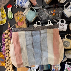 May include: Vintage striped handbag with a silver metal frame and chain handle. The fabric features vertical stripes in pink, blue, and cream. The bag is held up against a backdrop of various mugs and kitchen decor.
