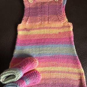 May include: A hand-knitted sleeveless pinafore dress featuring a horizontal striped pattern in pastel shades of pink, yellow, blue, and purple. The dress has two light-coloured wooden buttons at the shoulders and is paired with matching knitted booties.