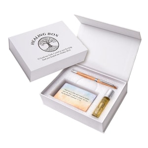 May include: A white rectangular gift box labeled HEALING BOX with a tree graphic and Psalm 30:2 text. Inside the foam-lined box is a light wood-toned pen, a small glass roller bottle with gold liquid, and a card featuring the text of Jeremiah 17:14.