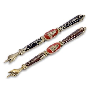 May include: Two metal Torah pointers, or yads, featuring a hand-shaped tip with an extended index finger. Each pointer has a central oval red enamel inlay with a gold-toned design, flanked by dark enamel sections with Hebrew and English text.