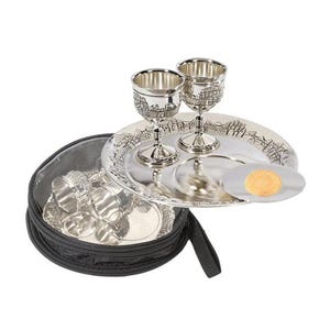 May include: A silver-toned metal set featuring two stemmed goblets and a matching round tray with embossed architectural details. A small circular disc with a tan center rests on the tray, and a black zippered storage case is included.