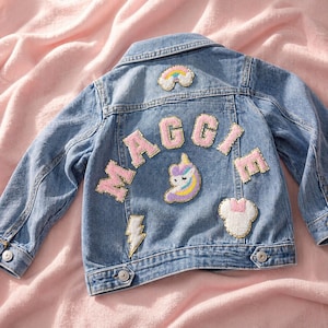 Puede incluir: Una chaqueta de mezclilla azul con el nombre MAGGIE en letras grandes de purpurina rosa y dorada en la espalda. La chaqueta está decorada con parches bordados que incluyen un arcoíris, una cabeza de unicornio, un rayo y una forma de oreja de ratón.