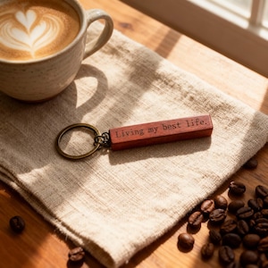 Puede incluir: Un llavero rectangular de madera con acabado marrón oscuro que presenta el texto grabado "Living my best life." unido a una anilla metálica de tono bronce. El llavero está colocado sobre una superficie de tela clara con textura.