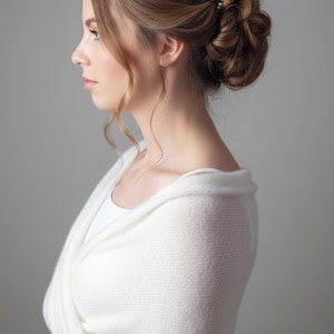 Op de afbeelding: Een vrouw met bruin haar in een knot, versierd met kleine parelhaarspelden. Ze draagt een witte sjaal over een witte top. De achtergrond is effen grijs.