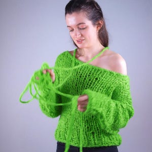 Puede incluir: Un suéter de punto grueso verde brillante con un diseño de hombros descubiertos. El suéter tiene mangas largas y un ajuste holgado. La modelo sostiene el hilo, mostrando la textura y el color de la prenda.