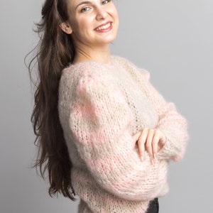 May include: A woman with long brown hair is wearing a pink and beige fuzzy sweater. She is smiling and has her arms crossed.
