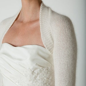 May include: A white knitted shrug with a scoop neck and long sleeves. The shrug is made of a soft, lightweight fabric and is perfect for layering over a dress or top.