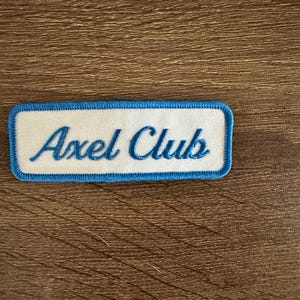 Peut inclure: Un écusson rectangulaire brodé présentant un fond blanc avec une bordure bleue. Le texte Axel Club est cousu en écriture cursive bleue au centre de l'écusson. Le design est net et présente une couleur de fil bleu uniforme et cohérente.