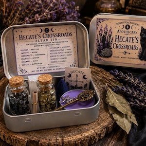 May include: A silver metal altar tin labeled Hecate's Crossroads containing two small glass jars with cork stoppers filled with dark stones and dried herbs, a small scroll, a purple disc, a metal key charm, and a card with a cross symbol.