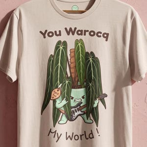 Peut inclure: Un t-shirt beige à manches courtes avec une illustration d'une plante verte en pot jouant de la guitare électrique. Le texte "You Warocq My World!" est imprimé en marron au-dessus et en dessous, avec une petite étiquette "Pork pour".
