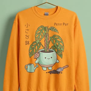 Peut inclure: Un sweat-shirt orange à col rond arborant une illustration d'une plante en pot vert clair souriante avec une feuille de monstera, un petit arrosoir et une pelle. Le texte "Petit Pot" et les caractères japonais "小さな鉢" sont imprimés au-dessus.