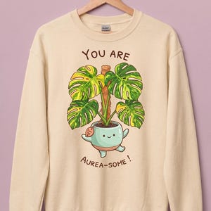 Peut inclure: Un sweat-shirt à col rond beige avec une illustration d'une plante en pot vert clair souriante, aux feuilles de monstera panachées grimpant sur un tuteur en mousse. Le texte au-dessus indique YOU ARE et en dessous AUREA-SOME! avec une étiquette.