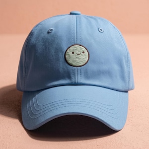 Peut inclure: Une casquette de baseball bleu clair avec un écusson brodé circulaire sur le devant. L'écusson est vert clair avec un contour marron foncé et représente un visage souriant simple avec deux petits points pour les yeux et une ligne courbe.
