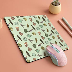 May include: A rectangular mouse pad with a light green background featuring a repeating pattern of gardening tools, potted plants, and small smiling faces. A light pink computer mouse with a delicate white floral line art design sits on the pad.