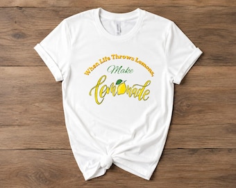 May include: A white short-sleeved t-shirt featuring the text When Life Throws Lemons, Make lemonade in yellow and green script and block lettering, with a small yellow lemon illustration replacing the letter o in the word lemonade.