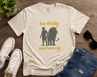 May include: A beige short-sleeve t-shirt featuring a dark gray silhouette of a person standing next to a lion. The text "Live Boldly" is printed in gold above the graphic, and the text "Lead Fearlessly" is printed in gold below the graphic.