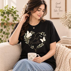 May include: A black short-sleeved t-shirt featuring a graphic of a heart-shaped wreath made of green leaves and white flowers. The design includes two white butterflies and the text Just Breathe written in a cursive script in the center.