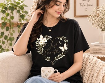May include: A black short-sleeved t-shirt featuring a graphic of a heart-shaped wreath made of green leaves and white flowers. The design includes two white butterflies and the text Just Breathe written in a cursive script in the center.