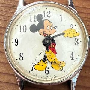 Peut inclure: Un cadran de montre-bracelet argenté illustré de Mickey Mouse avec des gants et des chaussures jaunes. Le cadran blanc affiche des chiffres arabes noirs de 1 à 12 et le texte WALT DISNEY PRODUCTIONS en bas. Le verre présente des rayures de surface.