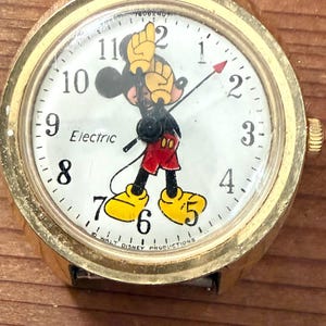 Könnte beinhalten: Eine goldfarbene Armbanduhr mit weißem Zifferblatt und einer Mickey-Maus-Grafik, deren Arme als Stunden- und Minutenzeiger dienen. Das Zifferblatt zeigt schwarze Ziffern, das Wort Electric und den Schriftzug Walt Disney Productions unten.
