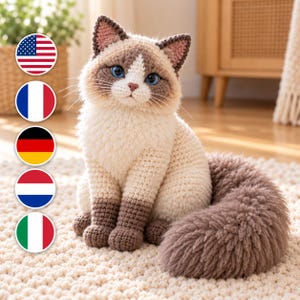 Könnte beinhalten: Ein gehäkeltes Stofftier in Form einer Katze mit cremefarbenem Fell am Körper, dunkelbraunen Pfoten, Ohren und Gesichtsmerkmalen sowie leuchtend blauen Augen. Die Katze hat einen flauschigen, strukturierten Schwanz und eine kleine rosa Nase.