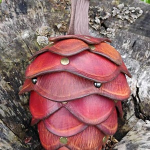 May include: A handcrafted leather pouch, shaped like a pinecone, in shades of red and brown. The pouch is adorned with brass rivets and a leather strap with a button closure. The item is displayed on a weathered tree stump.