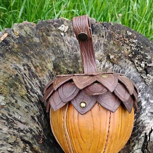 May include: A handcrafted leather acorn-shaped pouch in shades of brown and tan, with a leaf-like cap and a handle. The pouch is detailed with stitching and a brass rivet, set against a rustic wooden background.