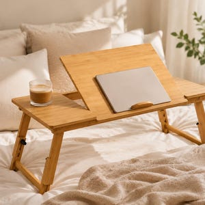 May include: A light brown bamboo laptop desk with an adjustable tilting surface, a small wooden ledge to hold a silver laptop, and two folding legs. A clear glass containing a light brown beverage sits on the flat side of the wooden desk.