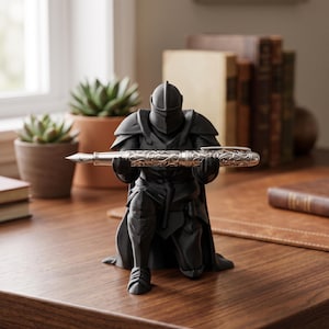 Peut inclure: Une figurine noir mat représentant un chevalier en armure complète agenouillé sur un genou. Le chevalier tient horizontalement devant sa poitrine un stylo plume argenté orné d'un motif floral gravé complexe entre ses mains.