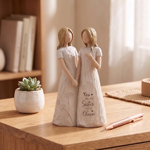 May include: A cream-colored resin figurine depicting two figures standing side-by-side, holding hands. The figures wear long, textured dresses. The front of the dress features the engraved text: You are the sister I got to choose, with small heart icons.